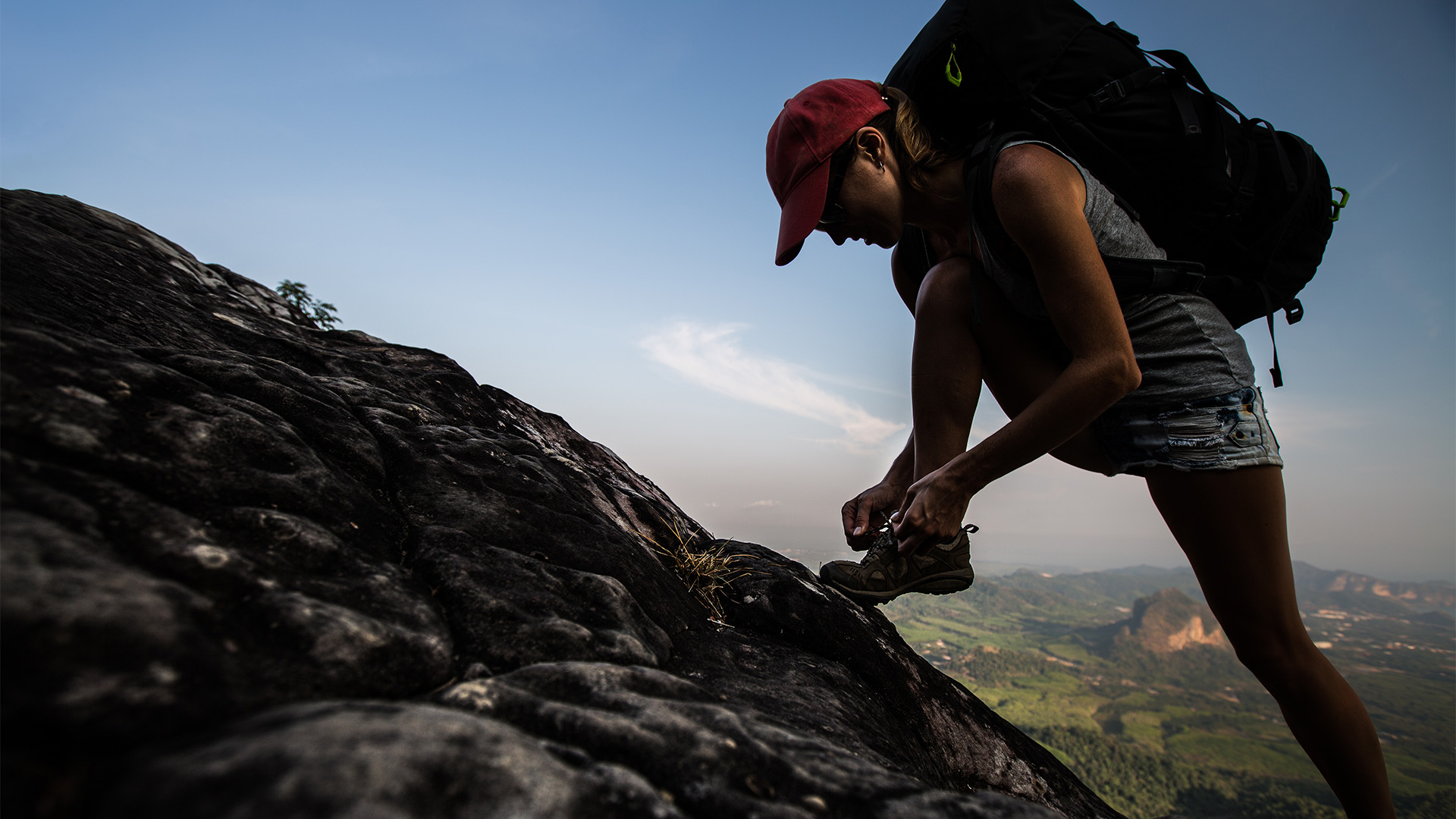 A female hiker planting her foot on a hillside and tying her hiking shoes, silhouetted against the countryside below