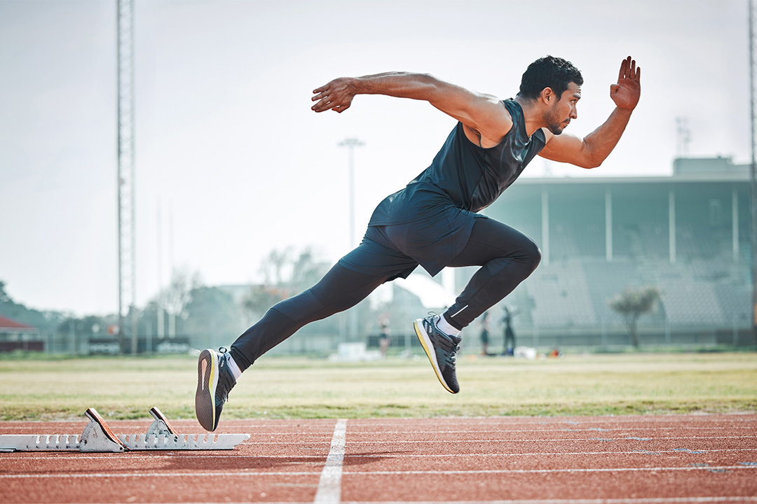 A man in a black tank top, black leggings, and black running shoes sprinting out of a set of starting blocks.